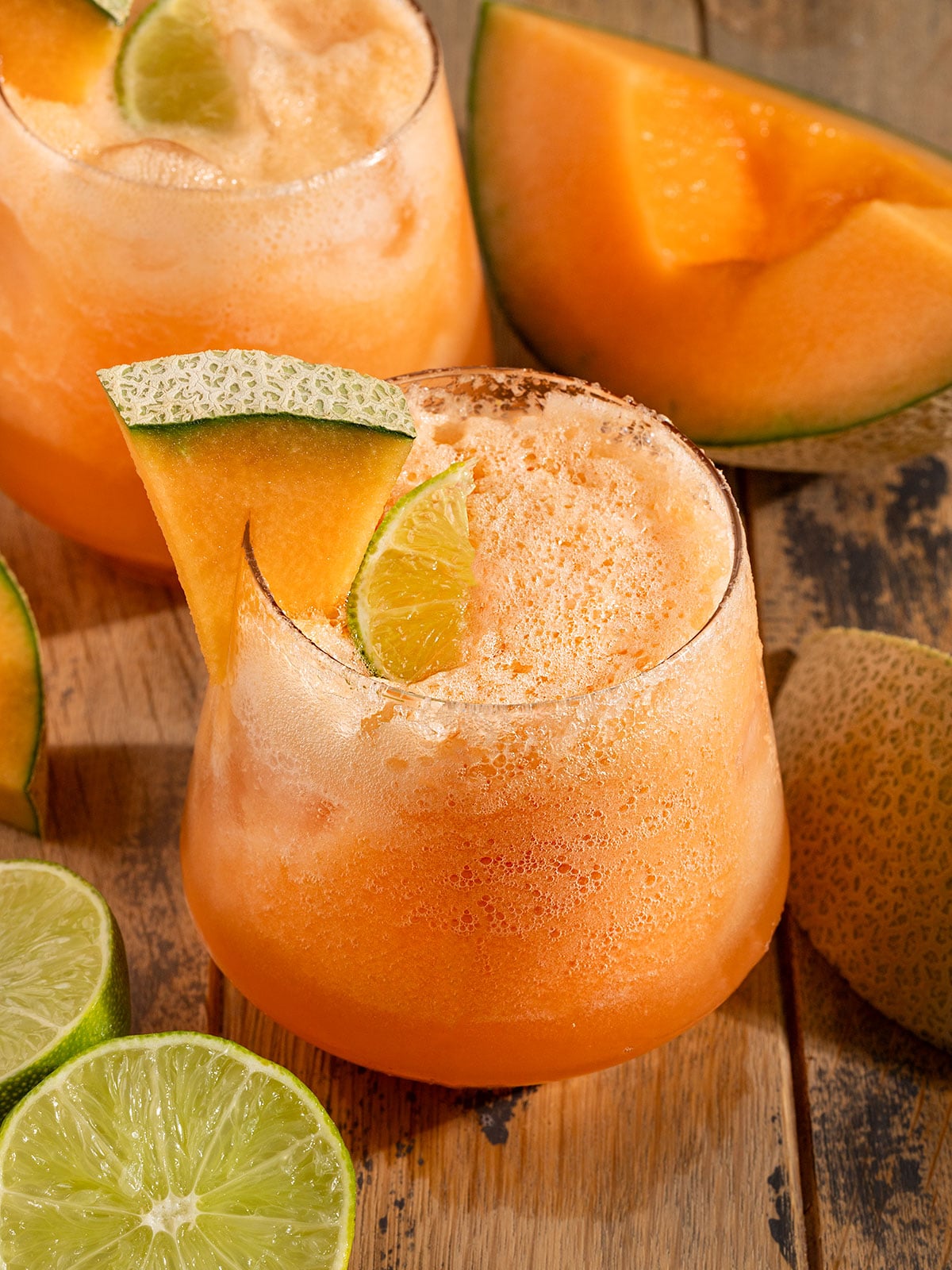 two glasses full of orange cantaloupe agua fresca with sparkling water and bubbles on top, a lime wedge, and a wedge of cantaloupe on the glass.