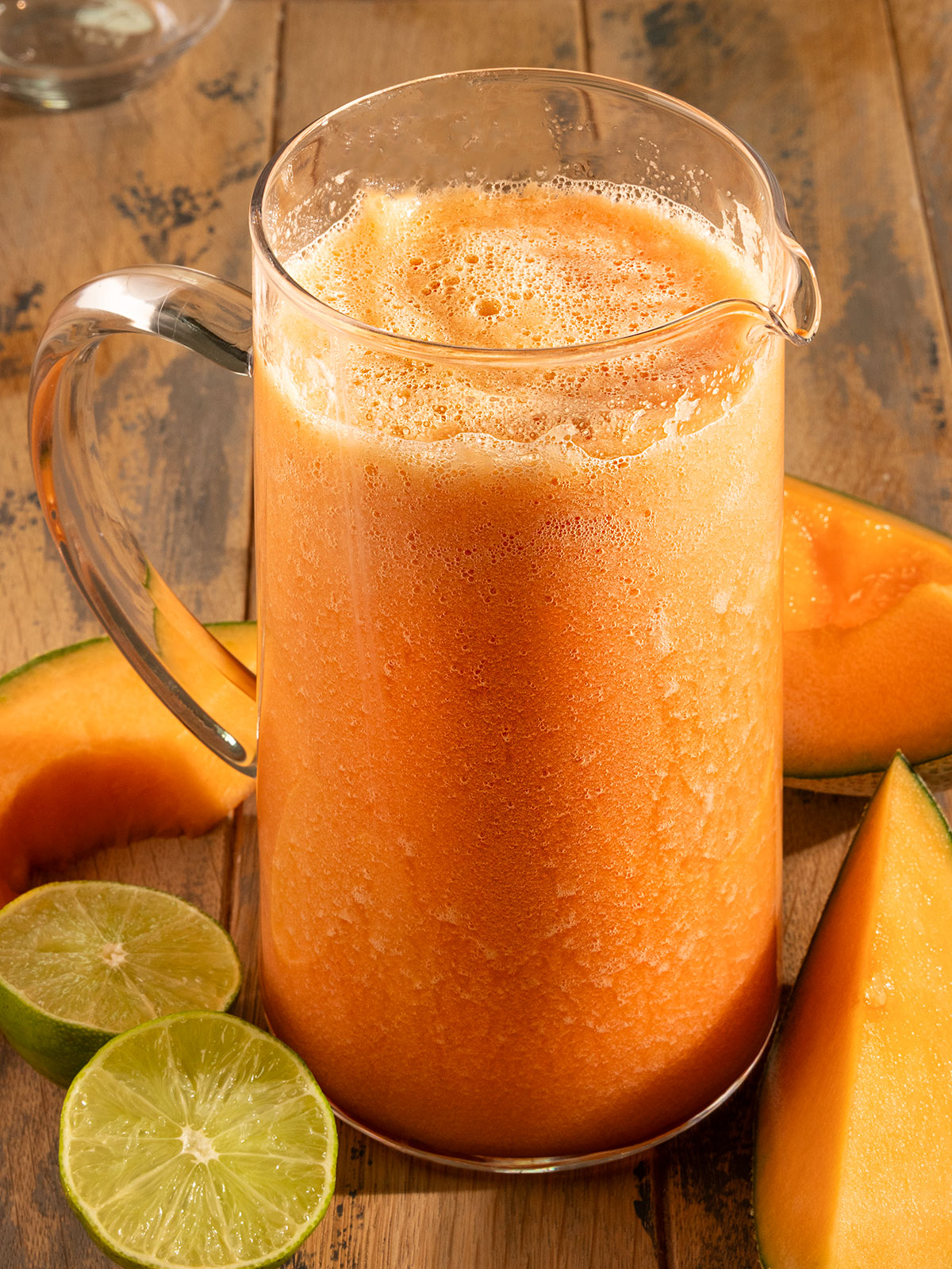 glass pitcher with orange cantaloupe juice inside surrounded by fresh limes and cantaloupe.