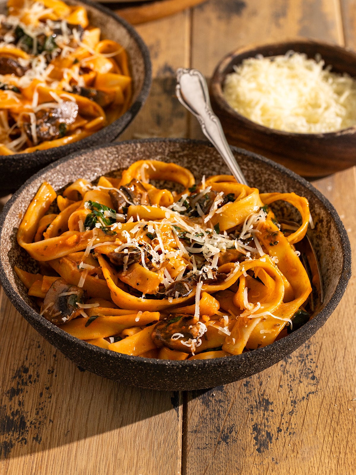 bowl of fettuccine with a red tomato sauce, mushrooms, spinach, and shredded parmesan cheese on top with fork in the bowl beside a bowl of shredded cheese and another bowl of pasta.