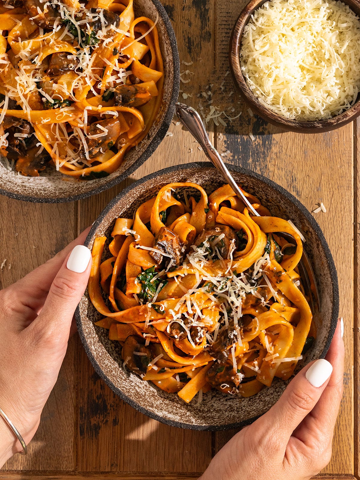 hand holding a bowl of  fettuccine pasta coated in a thick red tomato sauce with mushrooms, spinach, and parmesan cheese on top beside a bowl of shredded cheese and another bowl of pasta.