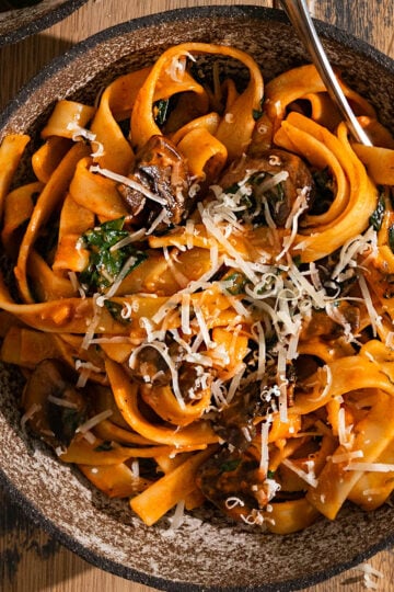 bowl of fettuccine with a red tomato sauce, mushrooms, spinach, and shredded parmesan cheese on top with fork in the bowl.
