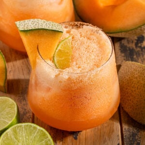 glass full of orange cantaloupe agua fresca with sparkling water and bubbles on top, a lime wedge, and a wedge of cantaloupe on the glass.