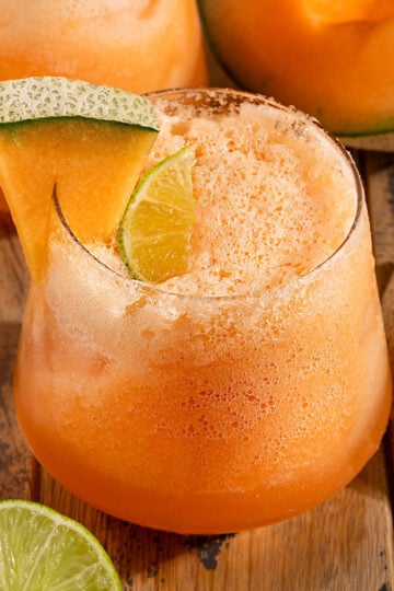 glass full of orange cantaloupe agua fresca with sparkling water and bubbles on top, a lime wedge, and a wedge of cantaloupe on the glass.