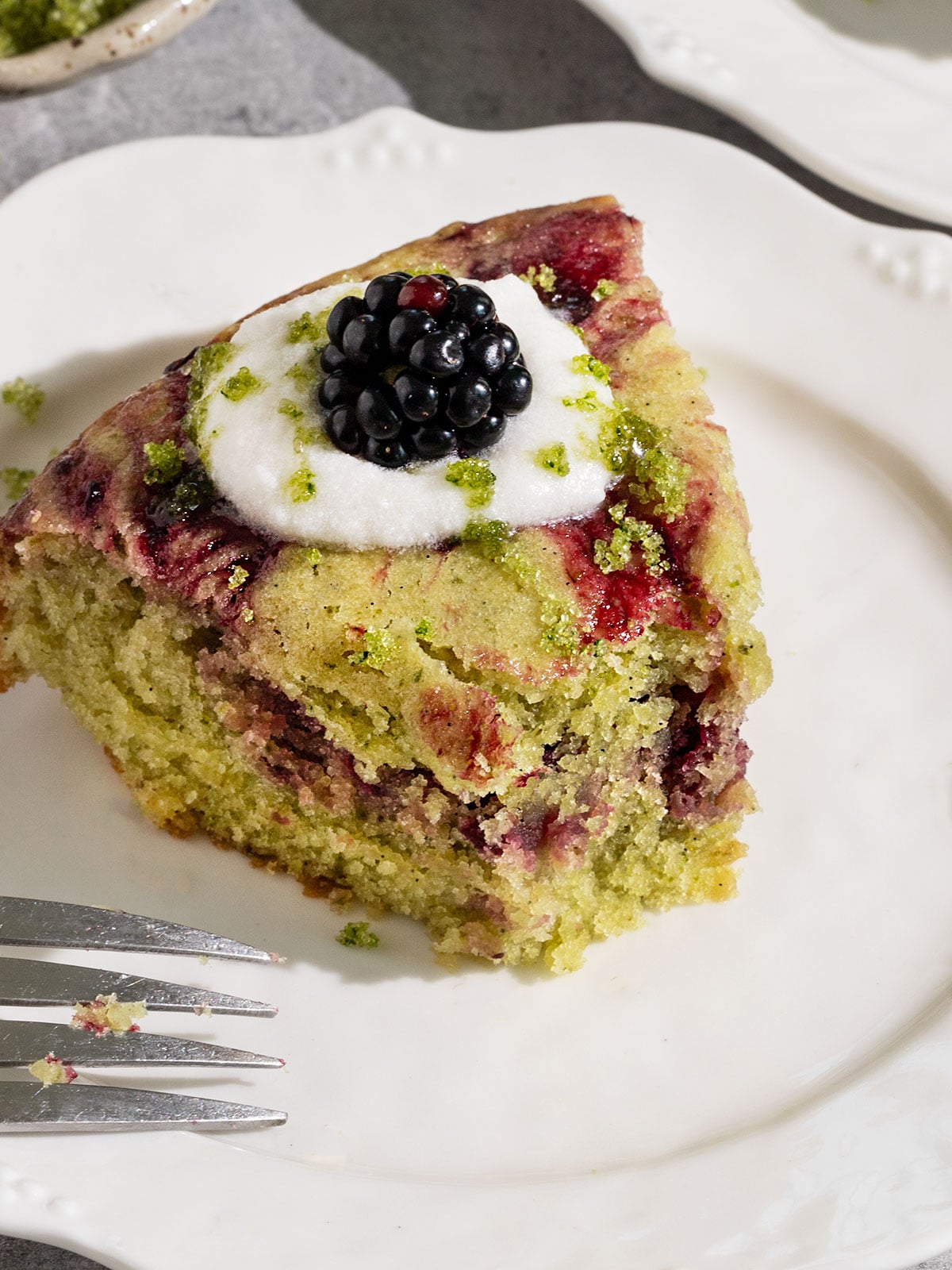 fluted plate topped with a slice of cake with swirls of blackberry jam topped with coconut cream, basil sugar, and fresh blackberries with a bite taken out.