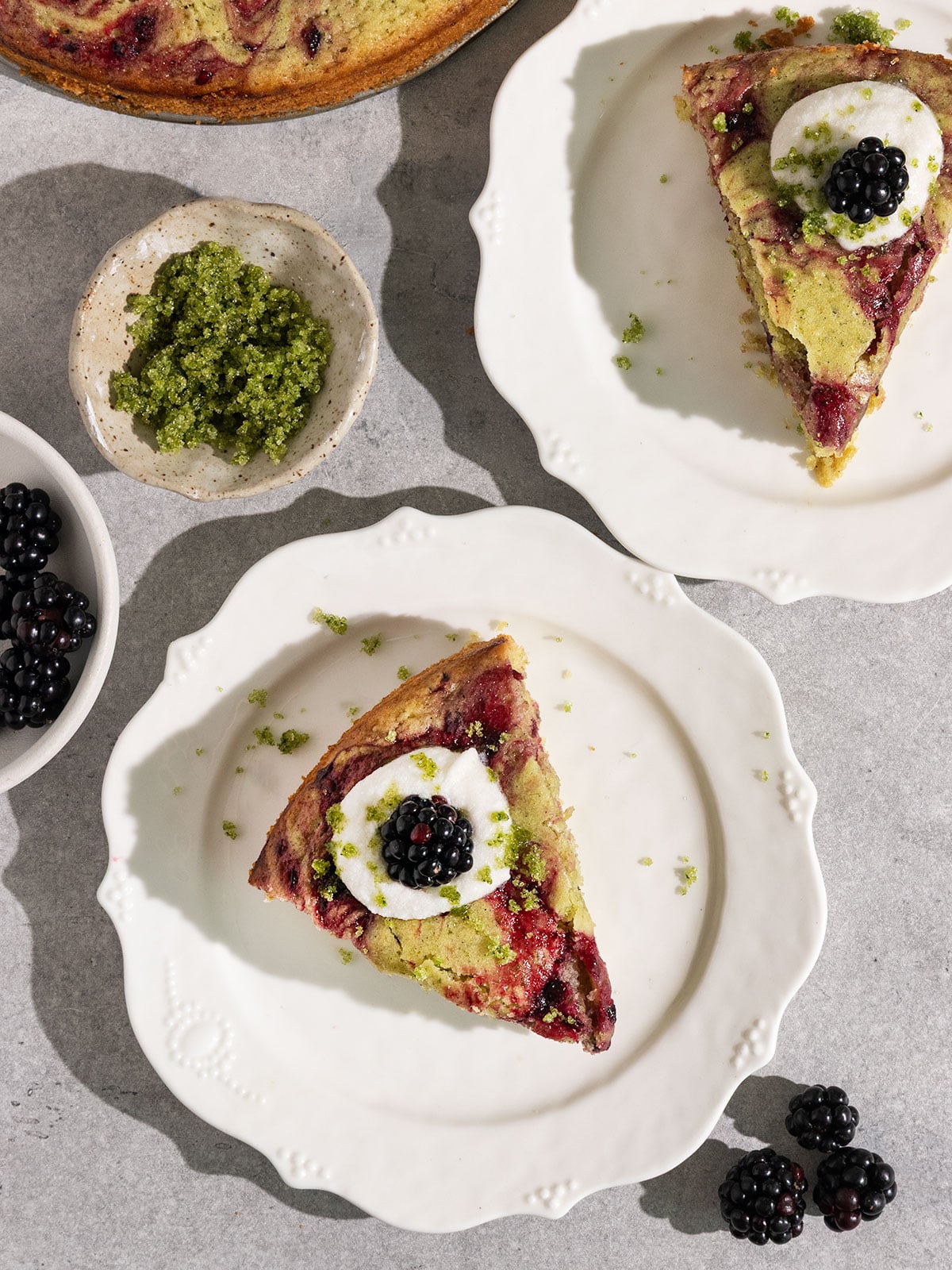 two fluted plates topped with slices of a green basil cake with swirls of blackberry jam topped with coconut cream, basil sugar, and fresh blackberries.