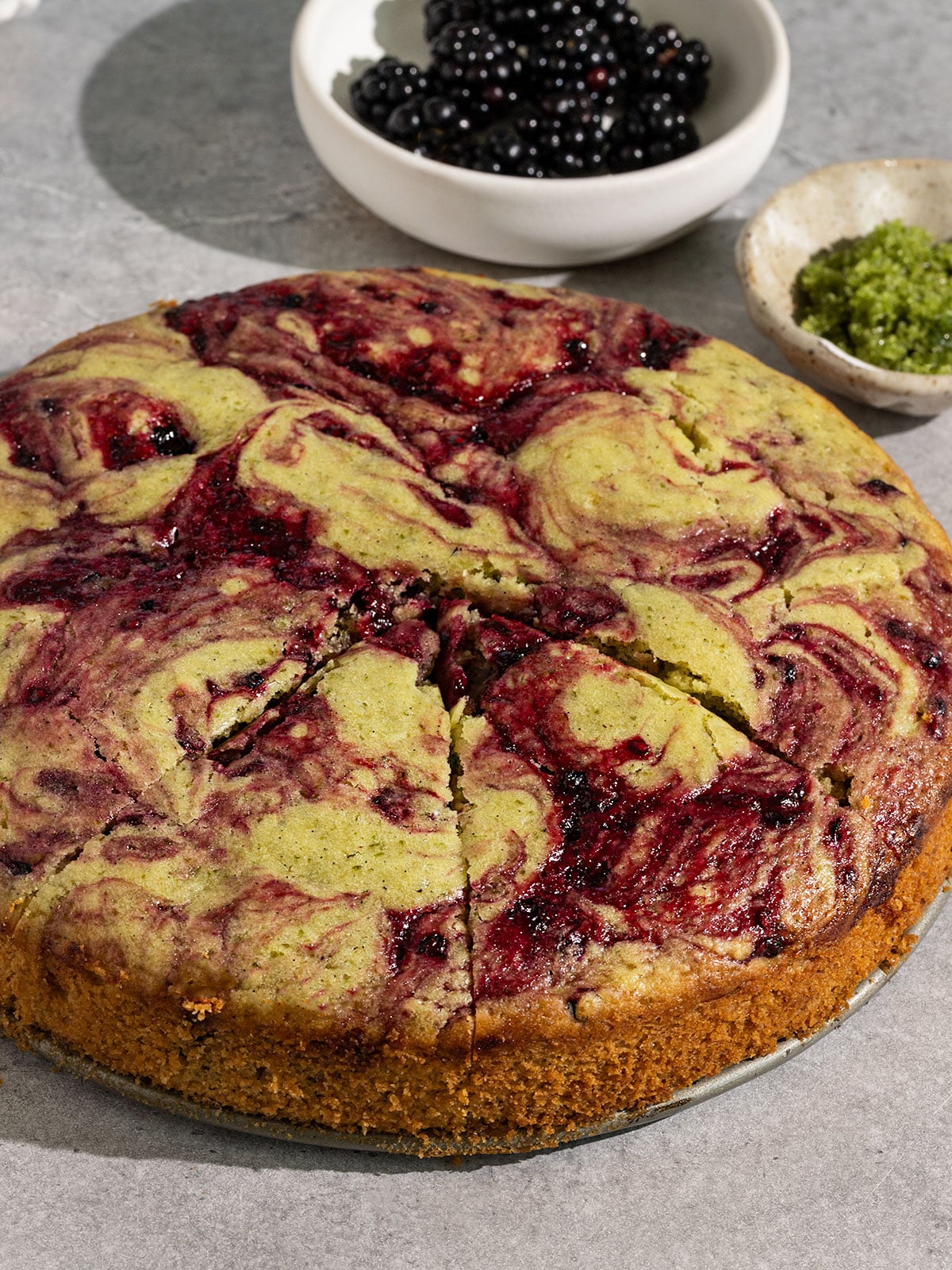 overhead view of a green basil cake with swirls of a purple pink blackberry jam with bowls of fresh blackberries and thai basil on side.