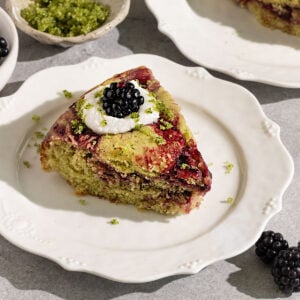 fluted plate topped with a slice of cake with swirls of blackberry jam topped with coconut cream, basil sugar, and fresh blackberries.