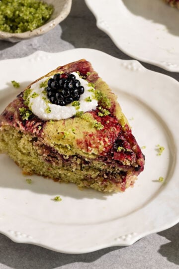 fluted plate topped with a slice of cake with swirls of blackberry jam topped with coconut cream, basil sugar, and fresh blackberries.