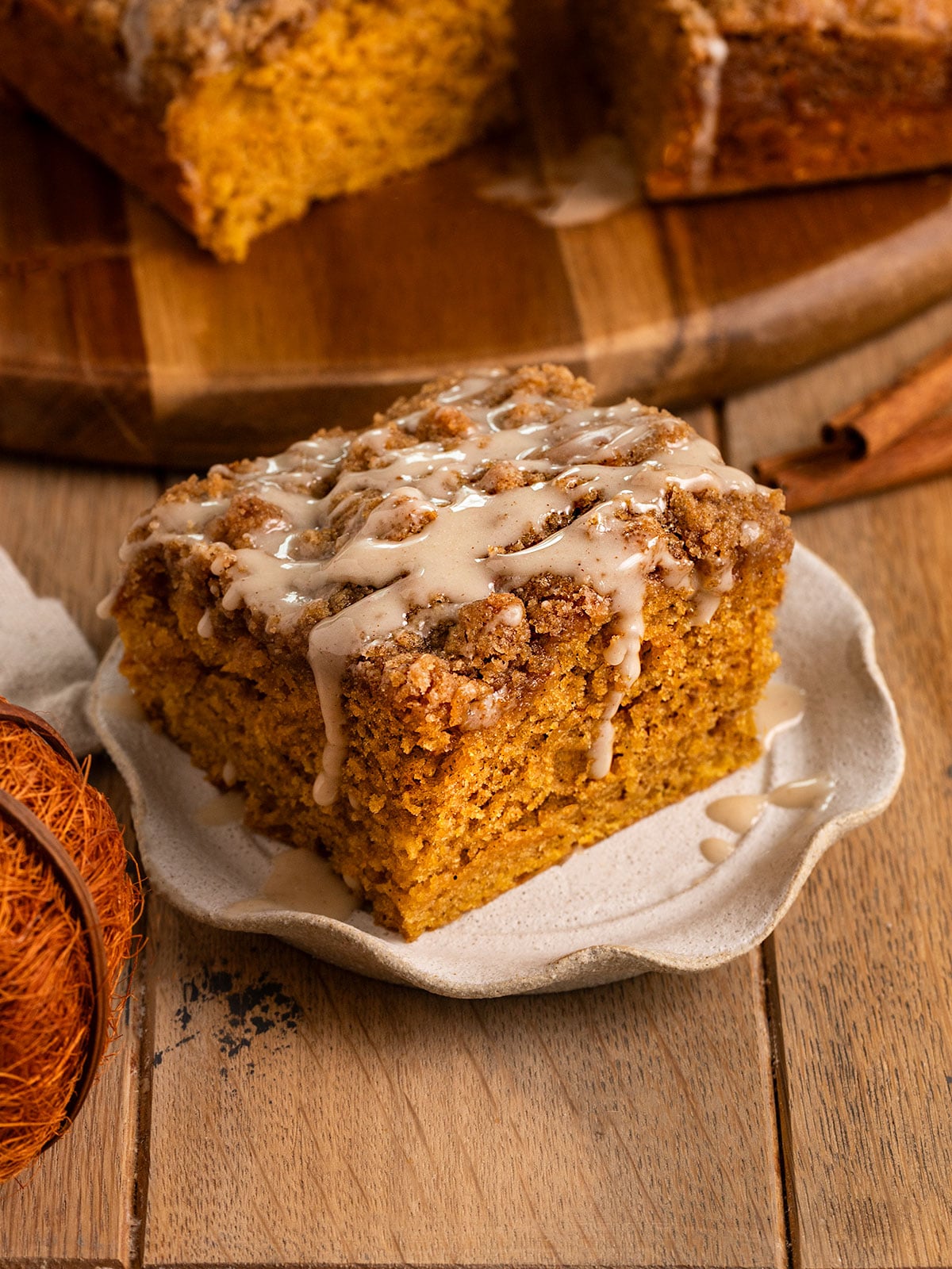 fluted plate topped with a slice of pumpkin cake topped with streusel and icing drizzled over the top in front of a board with more cake.
