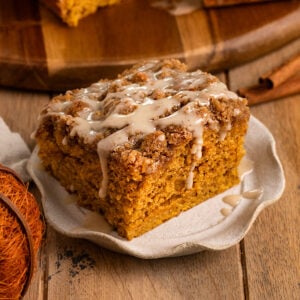 fluted plate topped with a slice of pumpkin cake topped with streusel and icing drizzled over the top in front of a board with more cake.