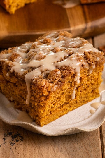 fluted plate topped with a slice of pumpkin cake topped with streusel and icing drizzled over the top in front of a board with more cake.