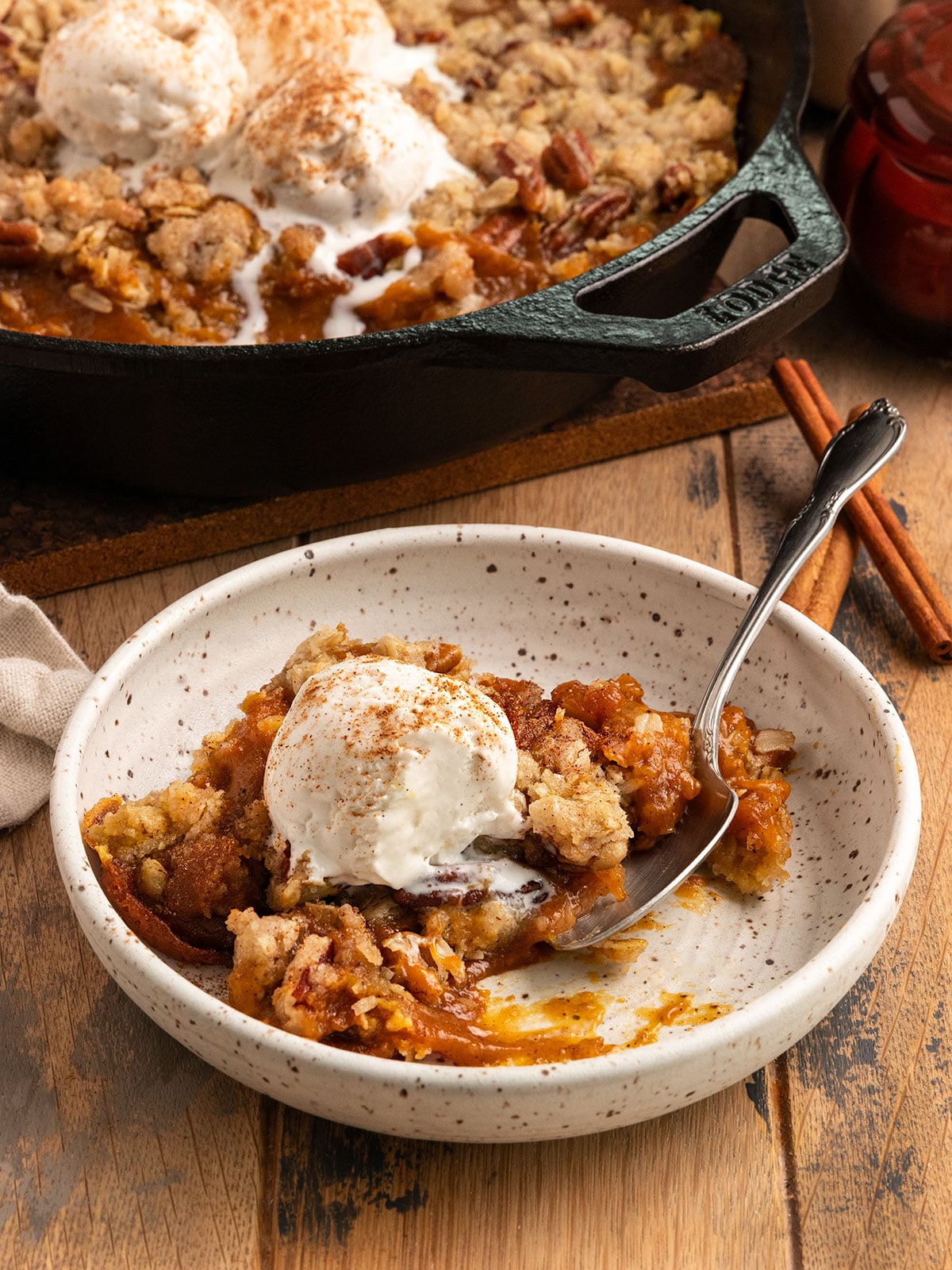 bowl of pumpkin pie filling with crisp topping and a scoop of ice cream on top and a spoon in the bowl alongside cast iron pan of more pumpkin crisp.