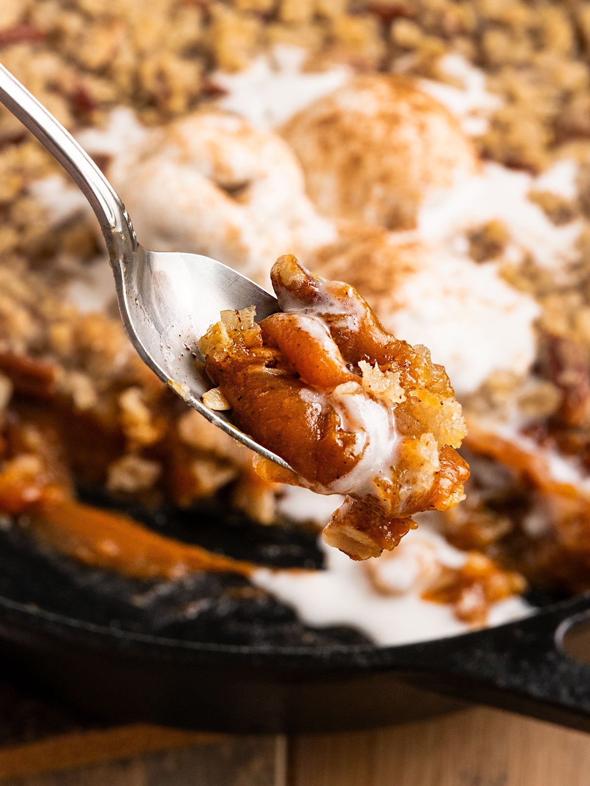 spoon holding scoop of pumpin filling topped with streusel crisp topping in front of skillet of more pumpkin pie crisp.
