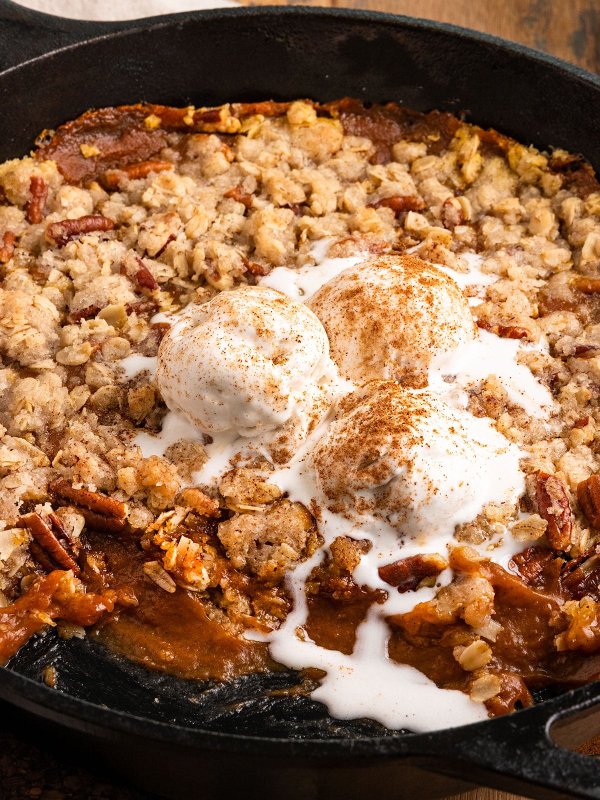 cast iron skillet full of a gooey orange pumpkin filling topped with a pecan streusel topping and scoops of melting vanilla ice cream.