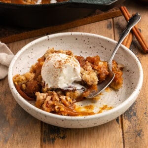 bowl of pumpkin pie filling with crisp topping and a scoop of ice cream on top and a spoon in the bowl alongside cast iron pan of more pumpkin crisp.