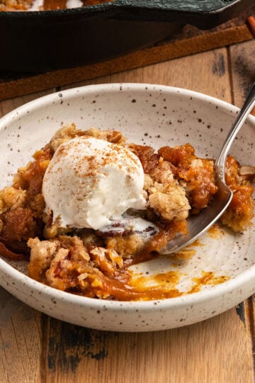 bowl of pumpkin pie filling with crisp topping and a scoop of ice cream on top and a spoon in the bowl alongside cast iron pan of more pumpkin crisp.
