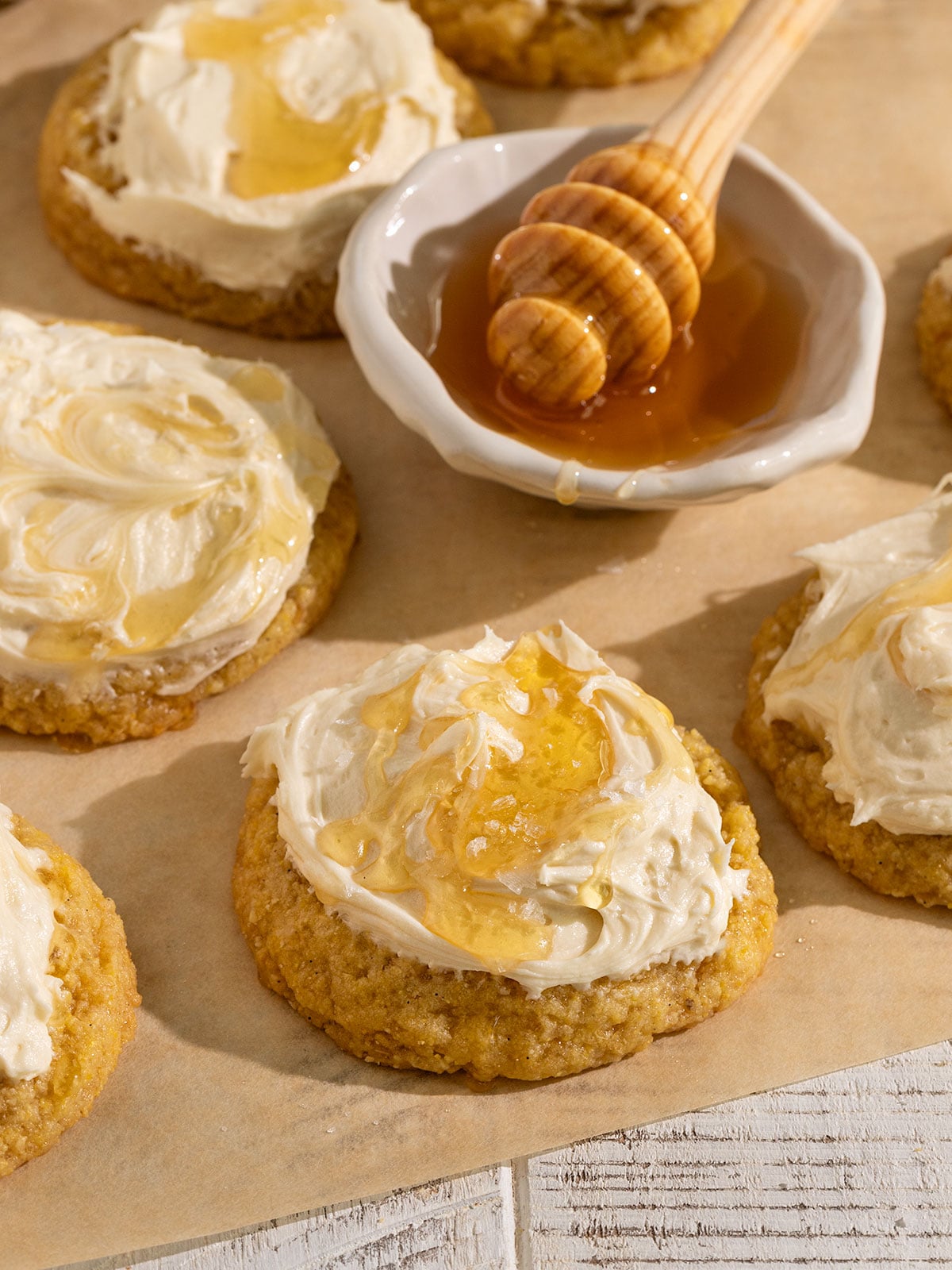 parchment paper topped with many yellow cornbread cookies topped with frosting, drizzle of honey, and flaky sea salt with a bowl of honey.