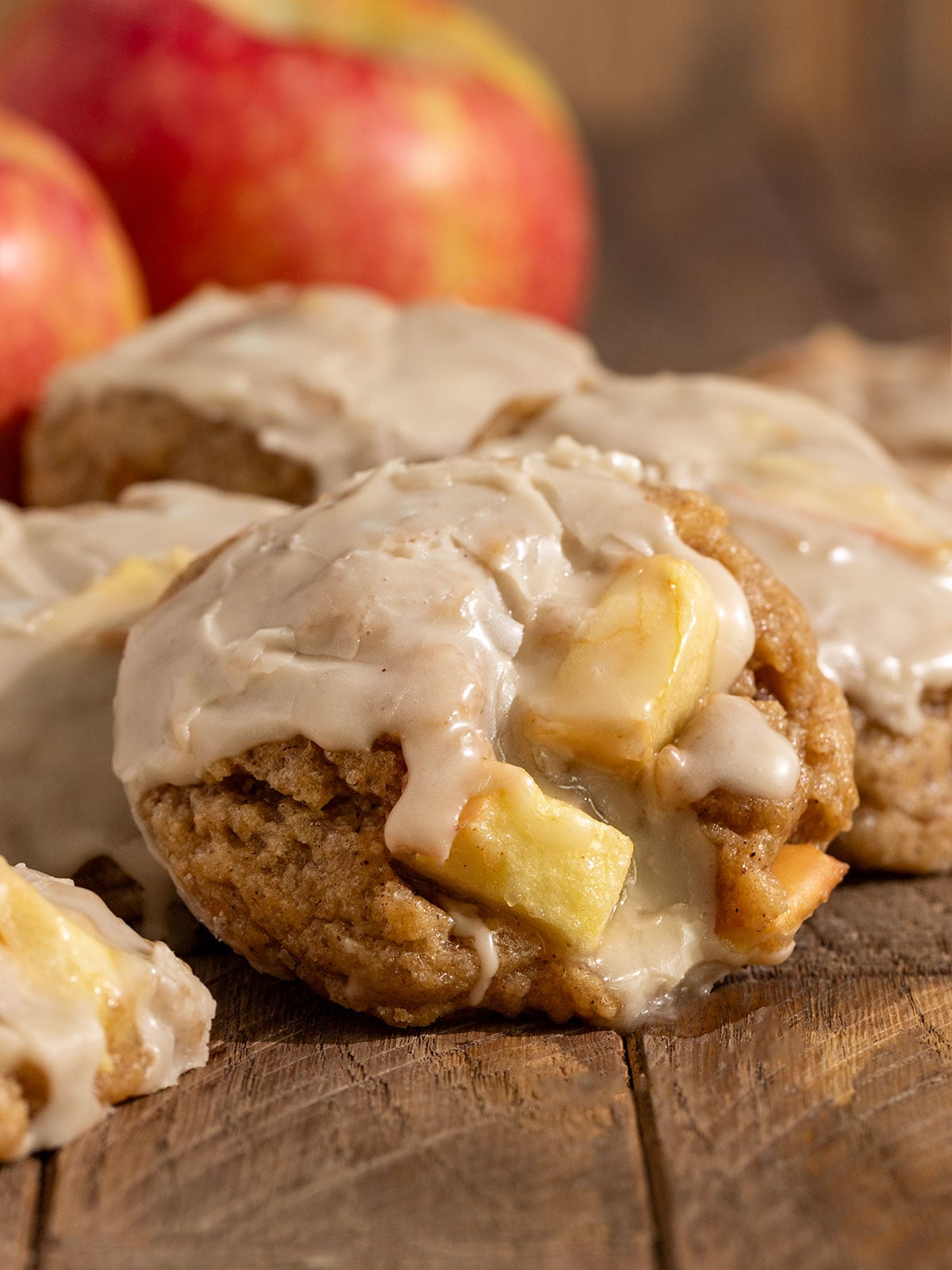 pile of cookies filled with apple chunks and topped with maple icing in front of fresh apples.