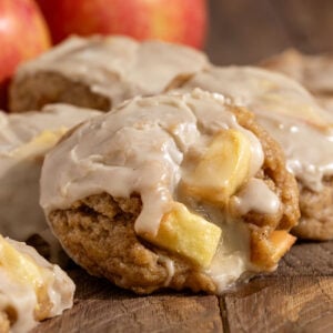 pile of cookies filled with apple chunks and topped with maple icing in front of fresh apples.