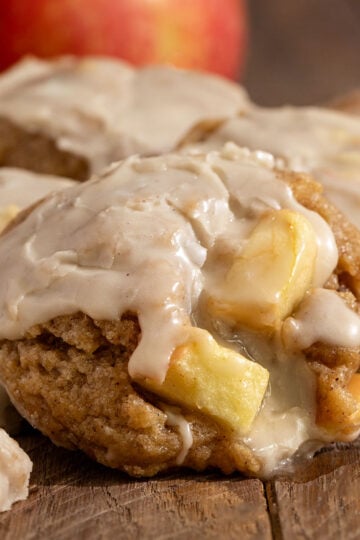 pile of cookies filled with apple chunks and topped with maple icing in front of fresh apples.