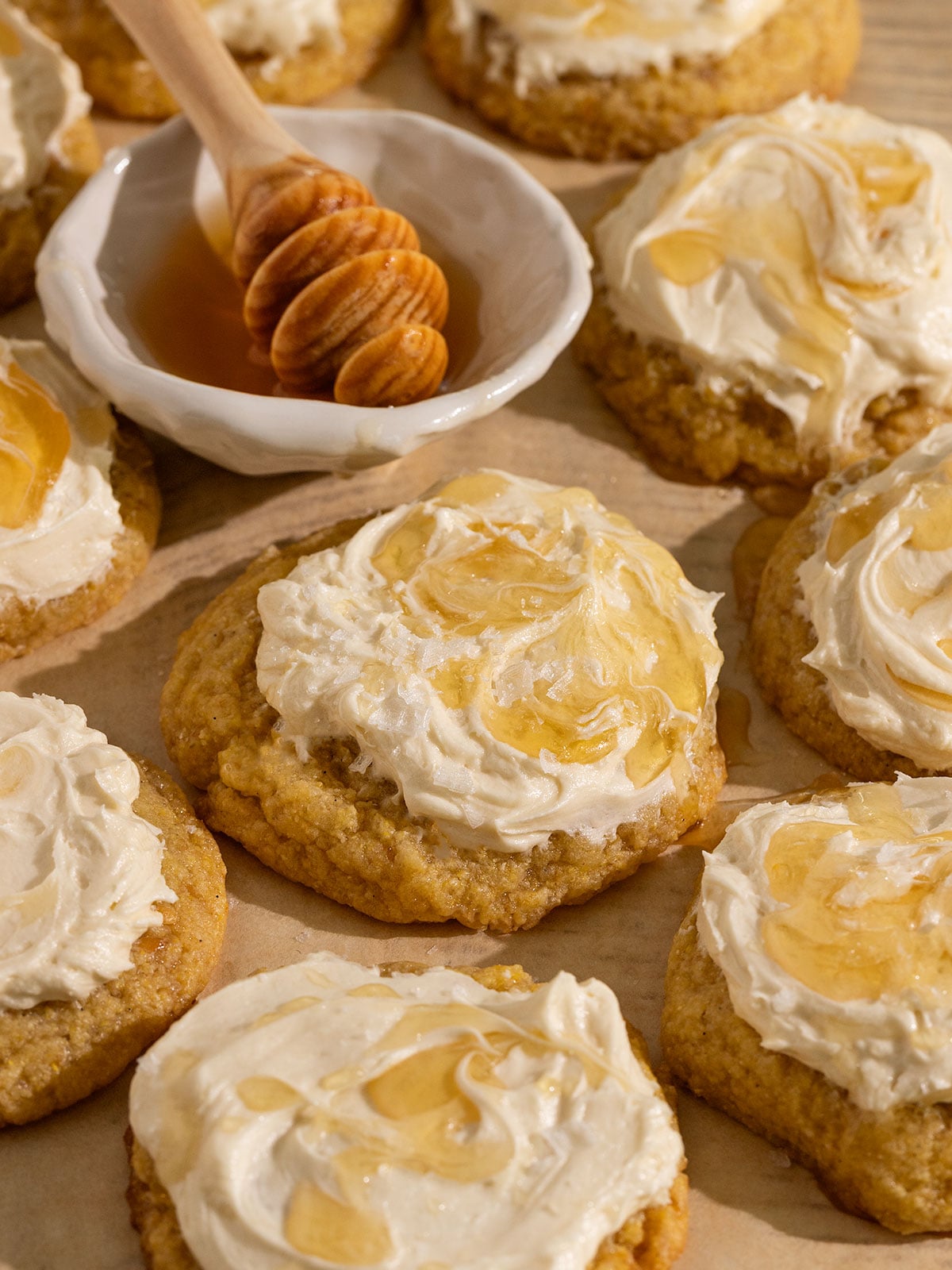 parchment paper topped with many yellow cornbread cookies topped with frosting, drizzle of honey, and flaky sea salt with a bowl of honey.