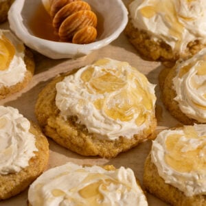 parchment paper topped with many yellow cornbread cookies topped with frosting, drizzle of honey, and flaky sea salt with a bowl of honey.
