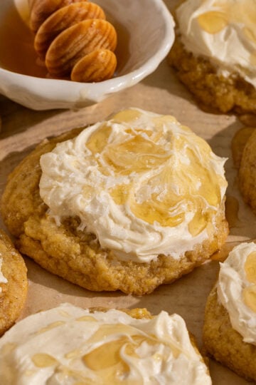 parchment paper topped with many yellow cornbread cookies topped with frosting, drizzle of honey, and flaky sea salt with a bowl of honey.