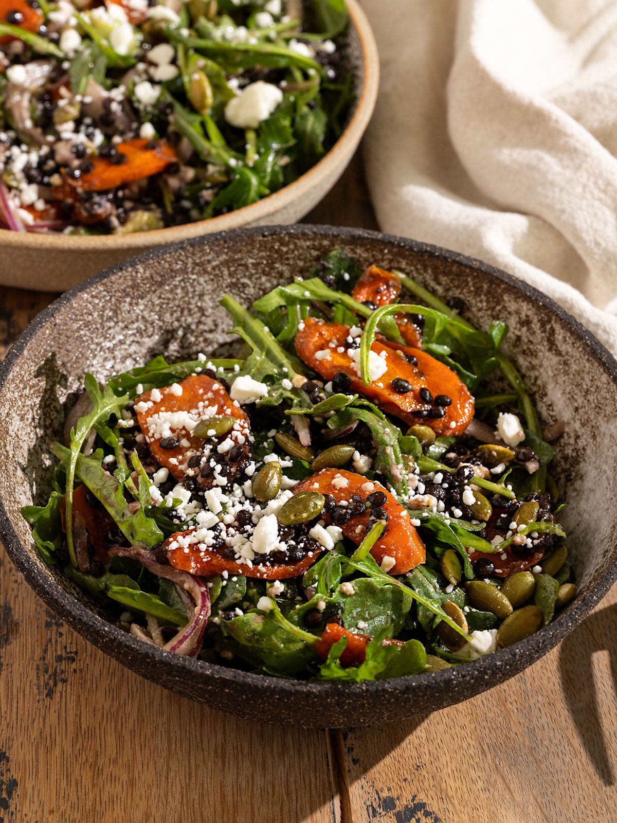 two bowls filled with a salad of arugula, roasted carrot slices, feta, red onion, avocado, pepitas, and black lentils topped with a dressing.