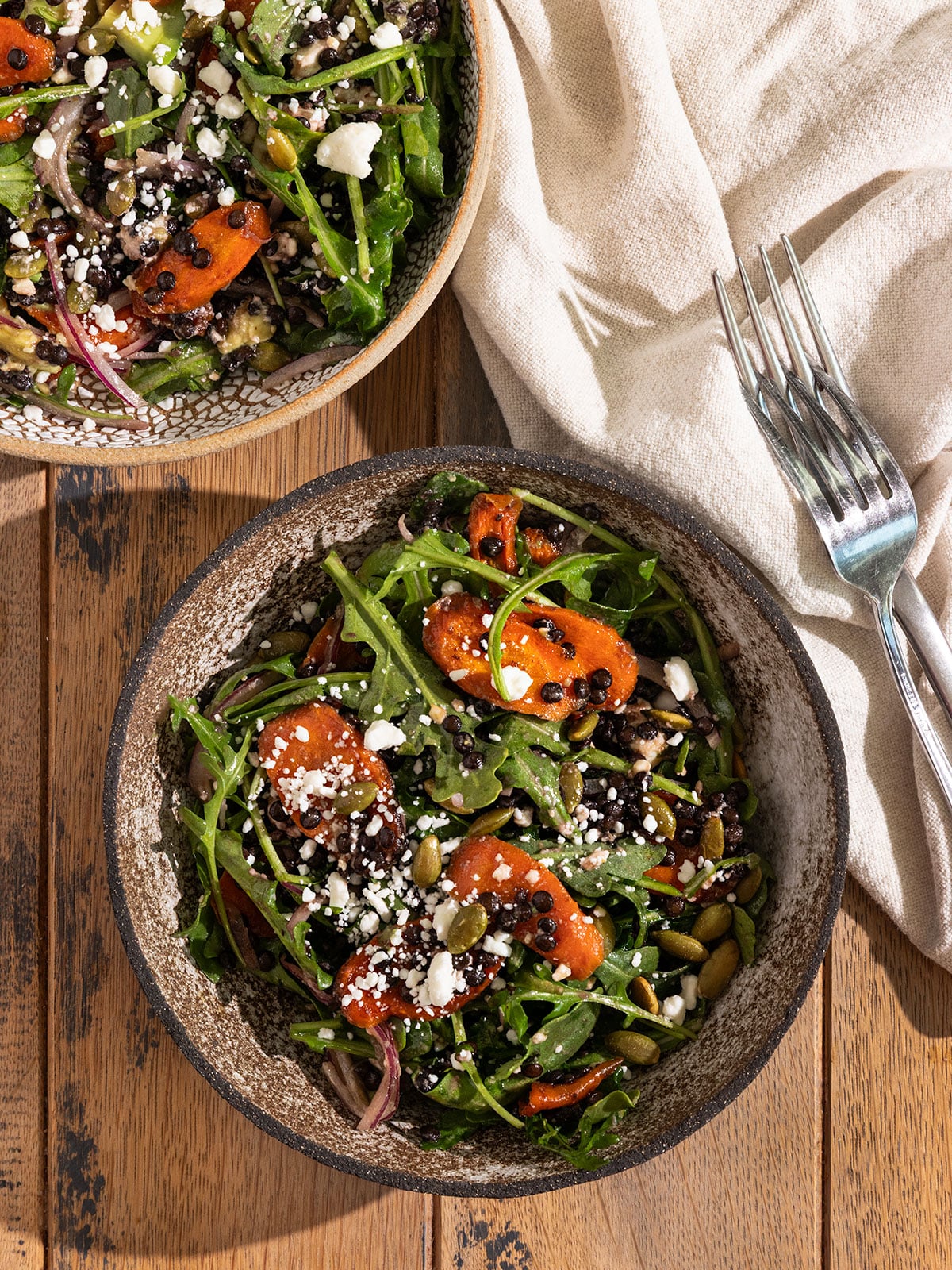two bowls filled with a salad of arugula, roasted carrot slices, feta, red onion, avocado, pepitas, and black lentils topped with a dressing and forks on the side.
