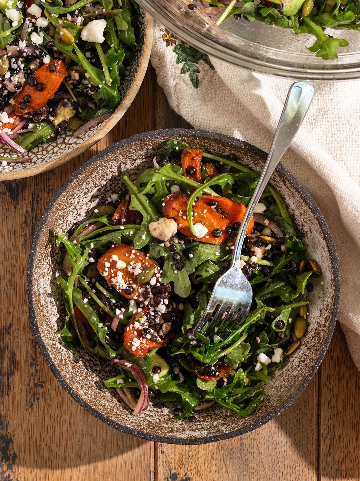 three bowls filled with a salad of arugula, roasted carrot slices, feta, red onion, avocado, pepitas, and black lentils topped with a dressing with front bowl having a fork in bowl.