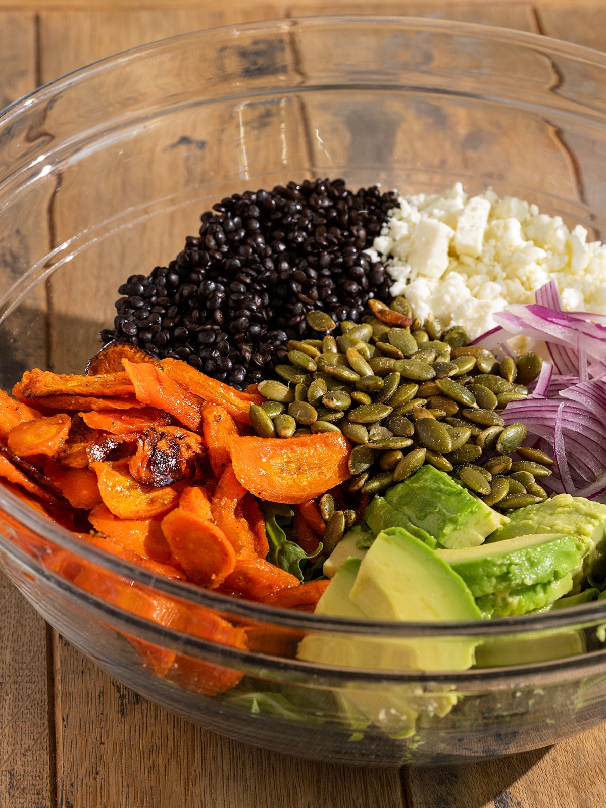 glass bowl filled with salad ingredients including arugula, roasted carrots, sliced red onion, crumbled feta, cooked black lentils, diced avocado, and roasted pepitas.