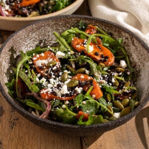 bowl filled with a salad of arugula, roasted carrot slices, feta, red onion, avocado, pepitas, and black lentils topped with a dressing.