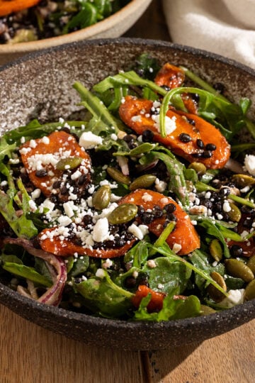 bowl filled with a salad of arugula, roasted carrot slices, feta, red onion, avocado, pepitas, and black lentils topped with a dressing.